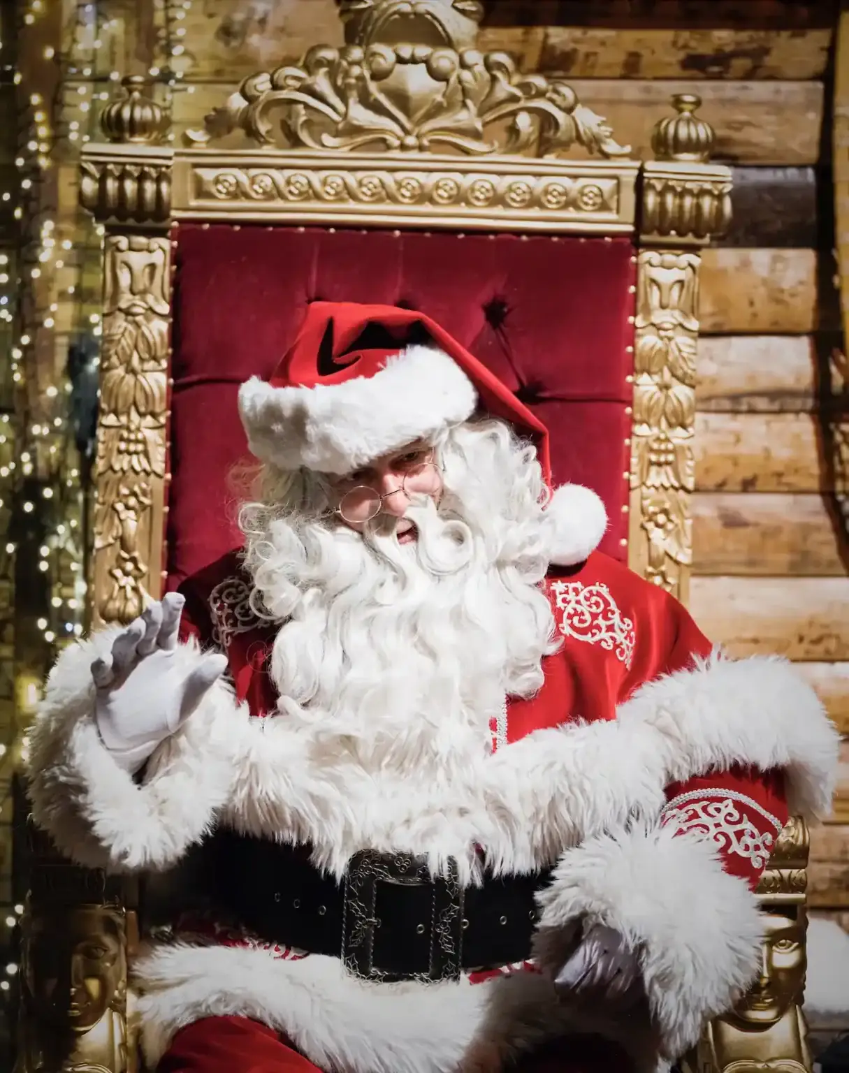 Le père Noël est assis sur son fauteuil devant un mur de chalet en bois